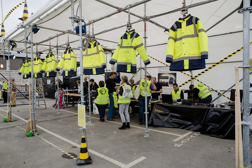 Série de vestes jaunes de chantiers suspendues sous un barnum sur un chantier, avec du public en arrière-plan 