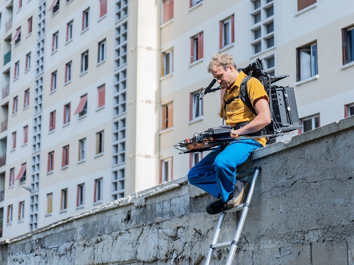 Un caméraman assis sur un muret devant une barre d’habitations dans le cadre du spectacle en déambulation « Impact d’une course » de la Compagnie La Horde à Bagneux en 2021
