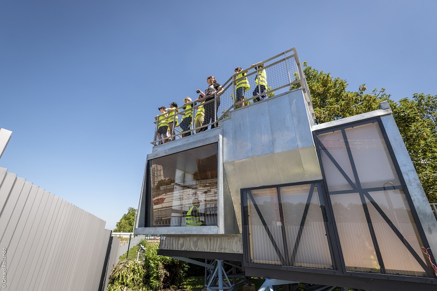 Belvédère en forme de maison des projets avec plusieurs enfants en gilets jaunes de chantier sur la plateforme, devant les palissades du chantier de Noisy-Champs
