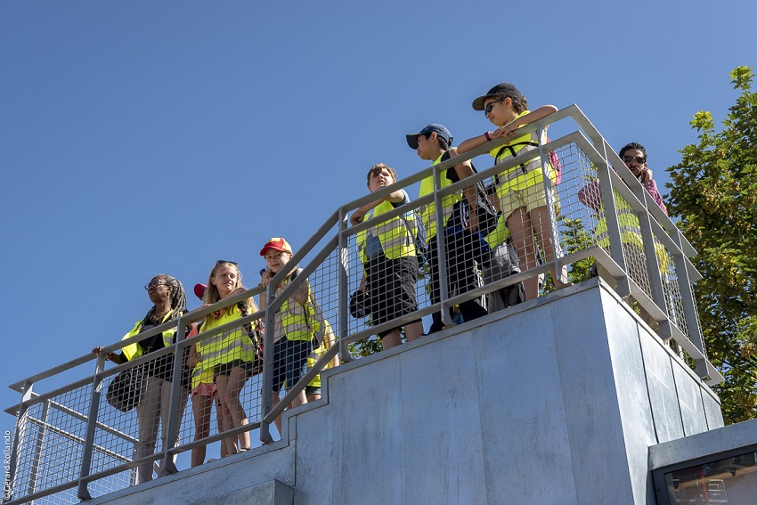 Belvédère en forme de maison des projets avec plusieurs enfants en gilets jaunes de chantier sur la plateforme, devant les palissades du chantier de Noisy-Champs