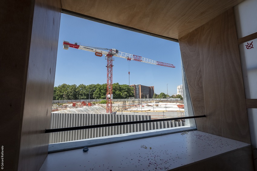 Intérieur du belvédère en forme de maison des projets avec plusieurs enfants en gilets jaunes de chantier sur la plateforme, avec vue sur la grue devant les palissades du chantier de Noisy-Champs