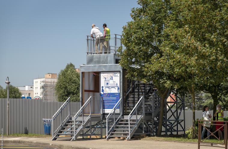 Belvédère en forme de maison des projets avec deux personnes sur la plateforme, devant les palissades du chantier de Noisy-Champs