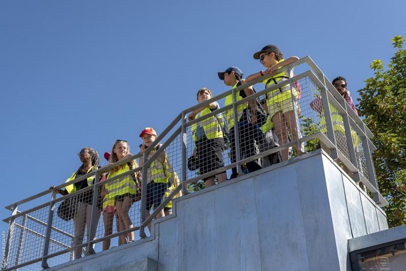 Belvédère en forme de maison des projets avec plusieurs enfants en gilets jaunes de chantier sur la plateforme, devant les palissades du chantier de Noisy-Champs