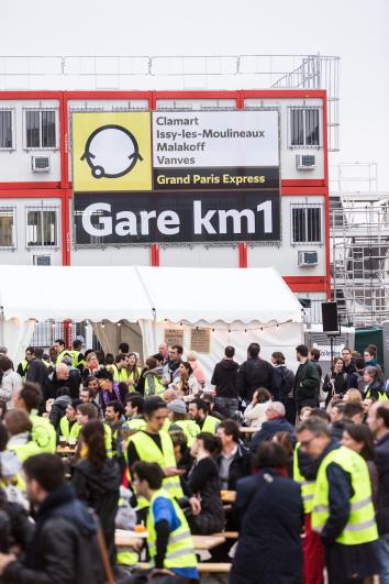 Public en gilet jaune de chantier, de jour, devant le panneau d’entrée du KM1 « Gare KM1 », sur le chantier de la future gare de Fort-d’Issy-Vanves-Clamart-Malakoff
