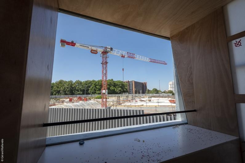 Intérieur du belvédère en forme de maison des projets avec plusieurs enfants en gilets jaunes de chantier sur la plateforme, avec vue sur la grue devant les palissades du chantier de Noisy-Champs