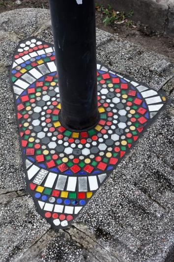 Motif avec des carreaux rouges, blancs, verts et jaunes Carreaux verts et blancs en cours de séchage sur un trottoir avec 4 plots orange de signalétique en arrière-plan, dans le cadre du projet « Flacking » par Ememem à Saint-Maur